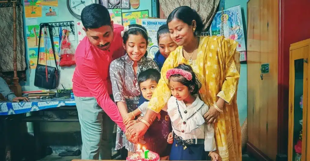 A family is gathered around a table, joining hands to cut a small birthday cake.