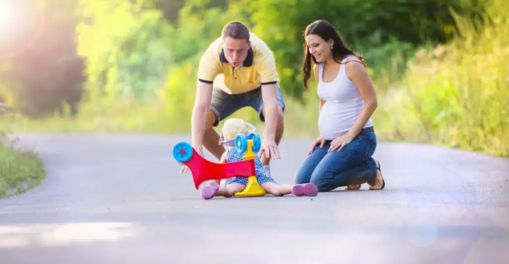 Parents assisting their toddler with a ride-on toy on an outdoor path.