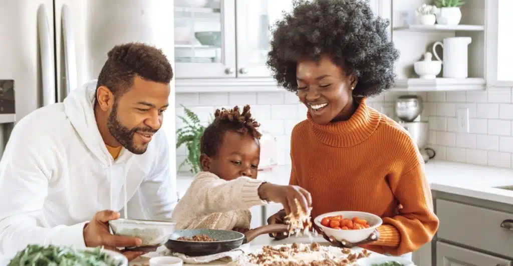 A family of three preparing and eating pizza together in a bright kitchen with the kid sprinkling cheese on the uncooked dough.