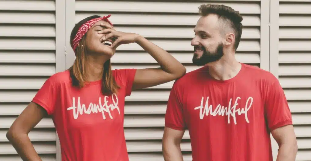 A couple stood in front of a wall wearing matching t-shirts and laughing while the woman touches her face .
