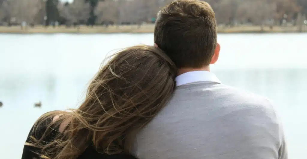 A couple sitting by a lake, with the woman resting her head on the man's shoulder.