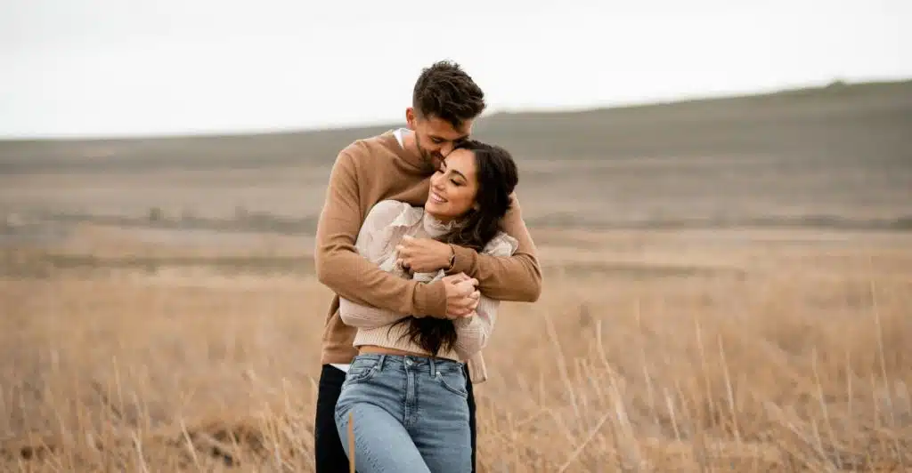 A man embracing a woman from behind in a field with a soft, overcast sky.