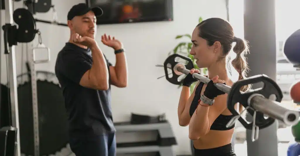 A trainer instructing a person lifting a barbell in a gym.