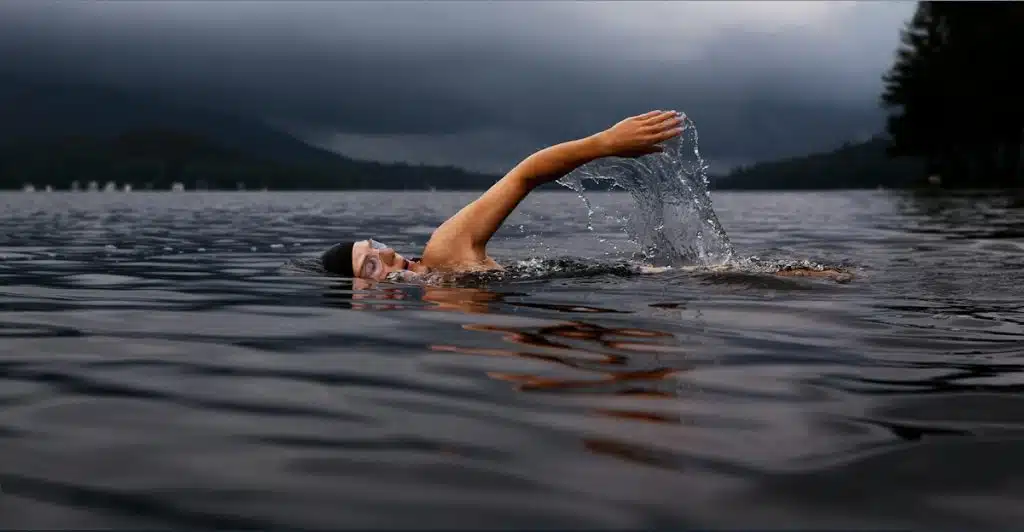 A person swimming in a dark lake with mountains and a stormy sky in the background.