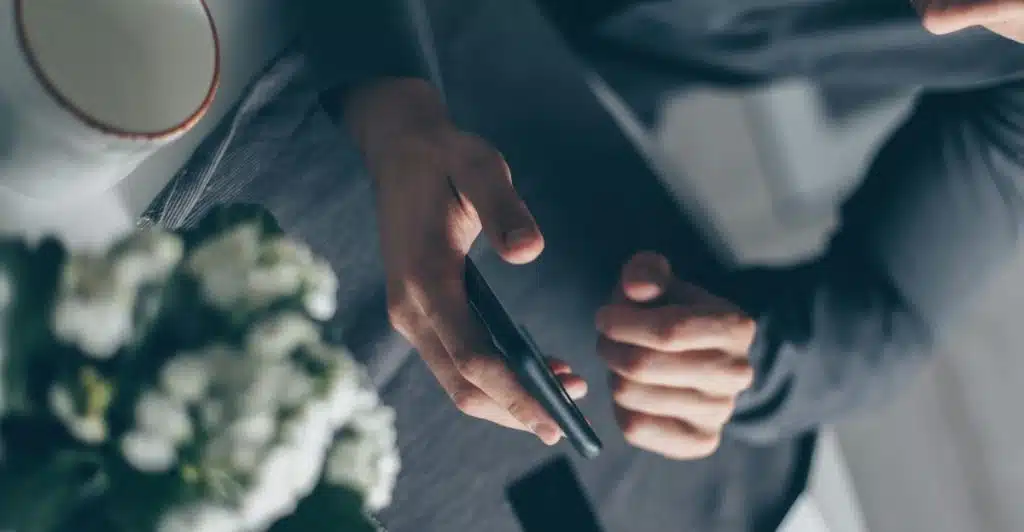 A man looking down at his phone, with papers, a card, and a partial view of a plant on a table.