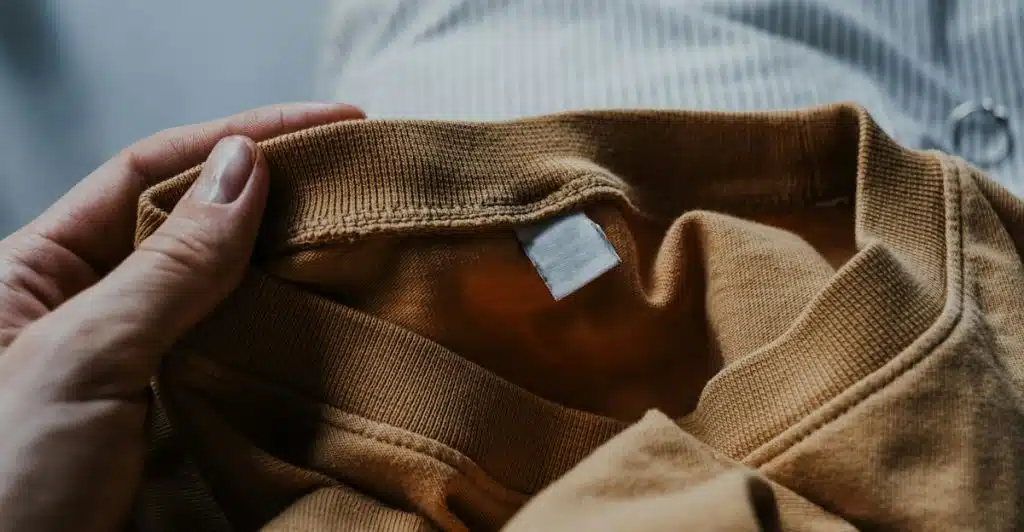 A hand holding a brown garment with a blank white tag and a striped fabric in the background.