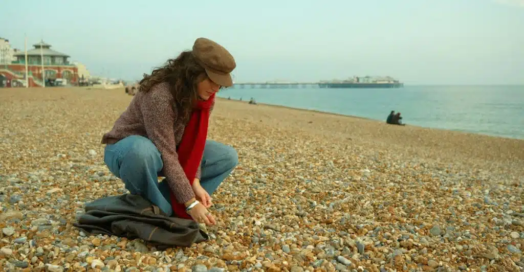 A person wearing a hat and a red scarf crouching on a rocky beach near the ocean.