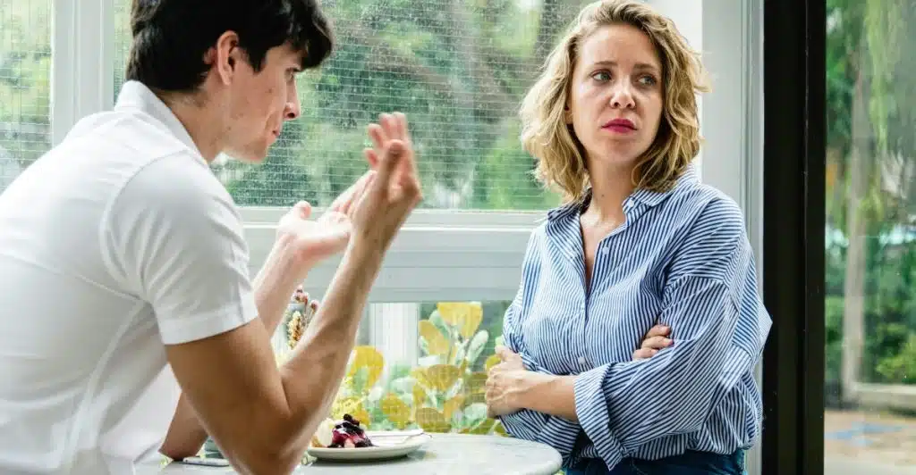 A man and a woman seated at a table in what appears to be a cafe or sunroom, engaged in a serious conversation.
