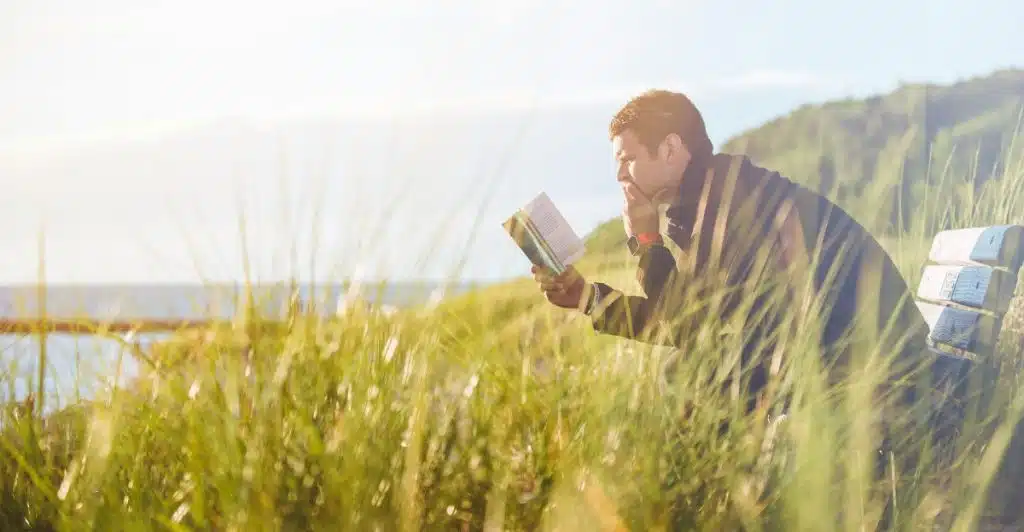 A person sitting on a bench outdoors, reading a book amidst tall grass.
