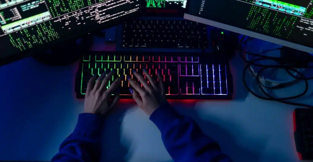 A person's hands typing on a brightly lit keyboard in front of computer screens.