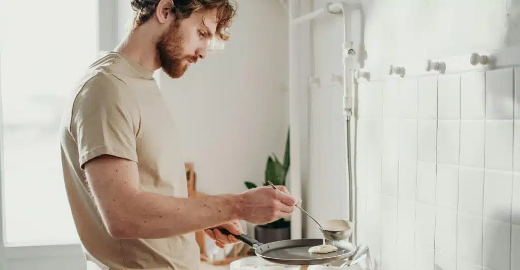 A person cooking pancakes in a pan on a stovetop in a kitchen.