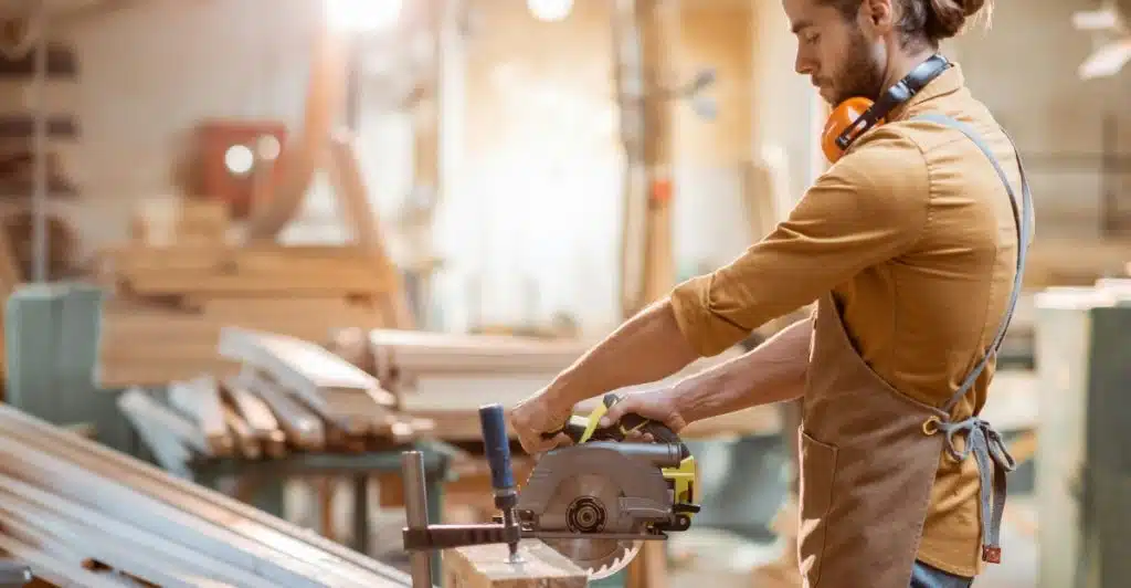 A person wearing safety gear using a circular saw to cut a piece of wood in a workshop.