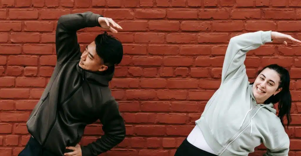 Two people in athletic wear stretching their arms in an outdoor setting with a brick wall behind them.