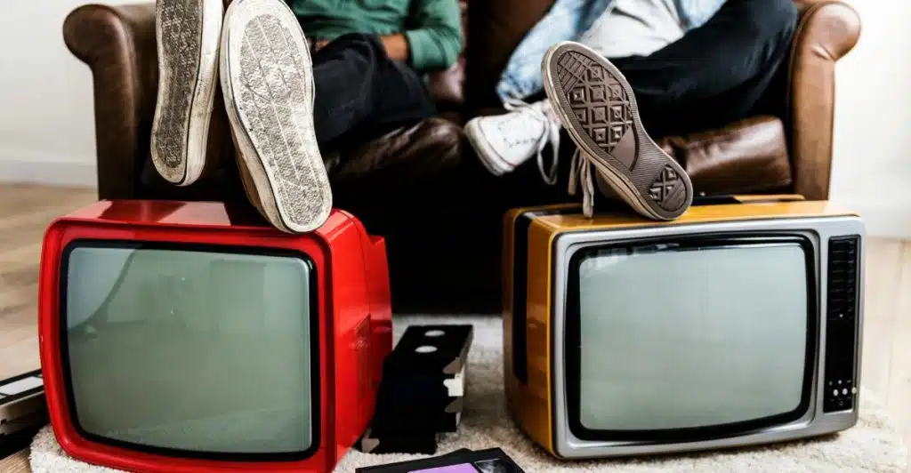 Two old-fashioned television sets and people sitting on a couch in the background with their feet on the TV sets.