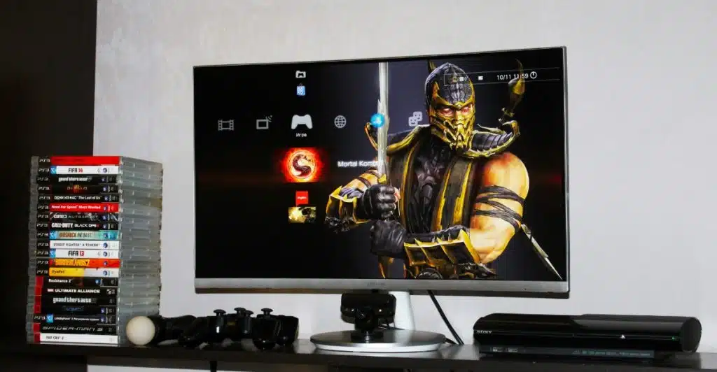 A computer monitor displaying a video game, with game controllers and a stack of game cases nearby.