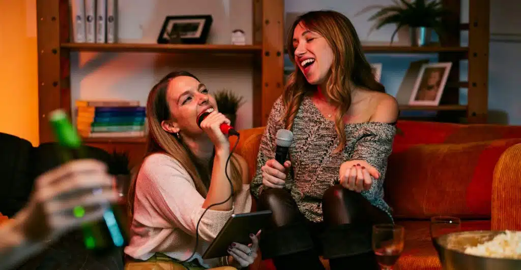 Two women singing karaoke on a couch with friends.