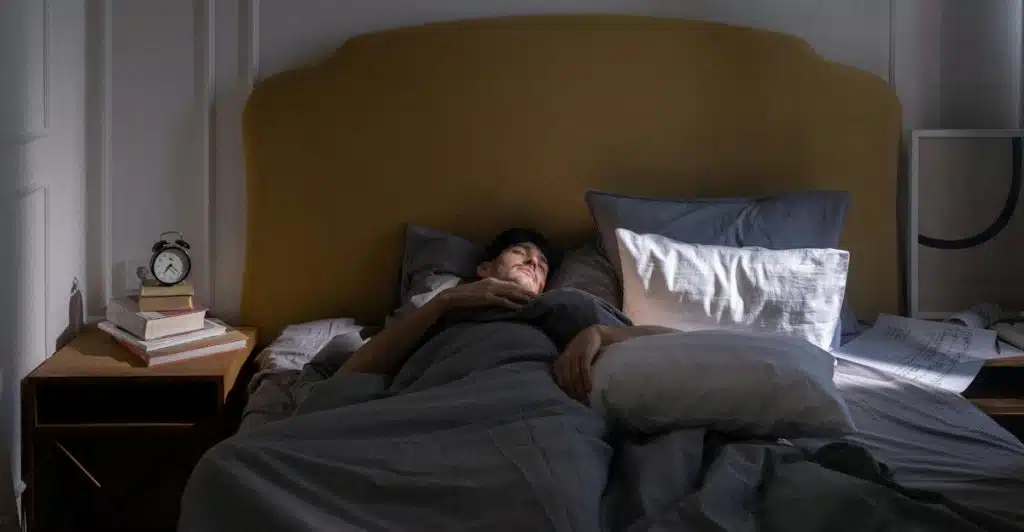 A man is lying in a bed with gray sheets, with an alarm clock and books on the nightstand.