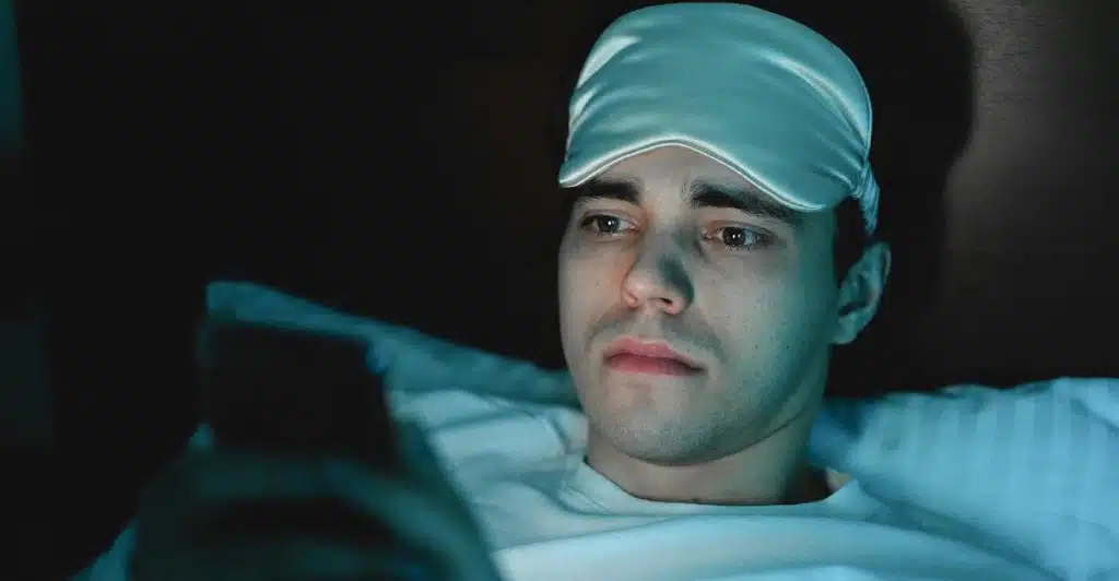 A man wearing a white sleep mask is looking at a phone in bed.