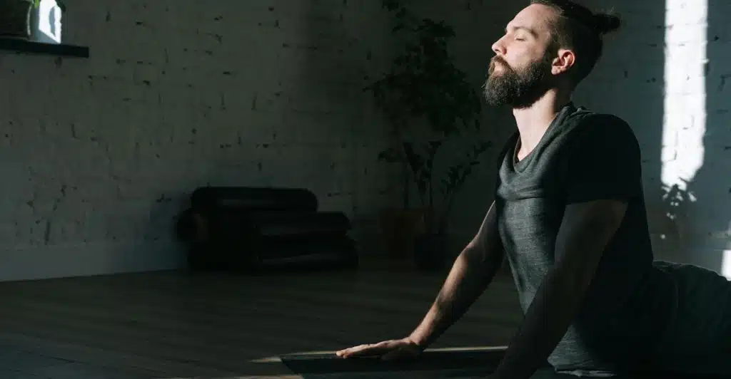 A man with a beard is doing a yoga pose on a mat in a room with sunlight.