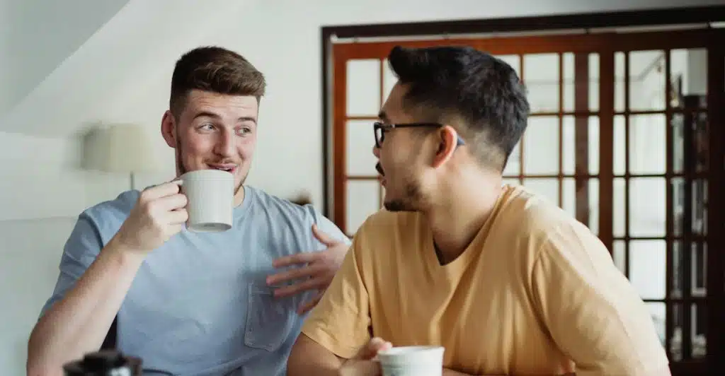 Two people are sitting at a table, drinking from mugs and talking.