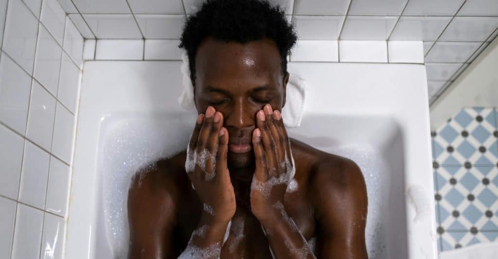 A man is washing his face with soapy hands in a bathtub.