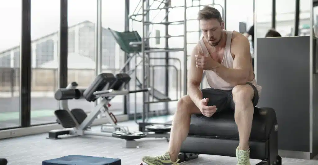 A man in a tank top and shorts sitting at a gym, looking tired.