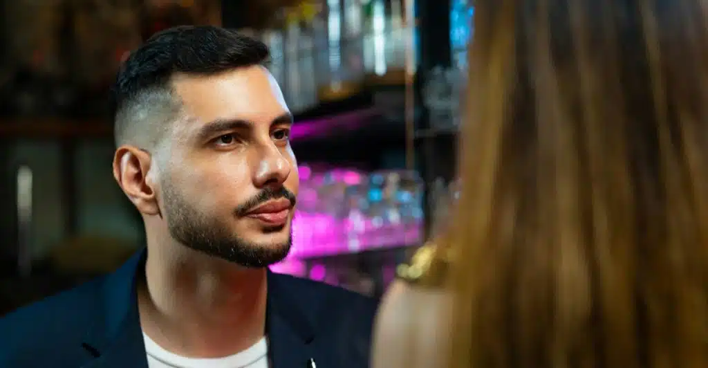 A man with a beard and a blue jacket looking intently at a woman with long brown hair in a bar setting.