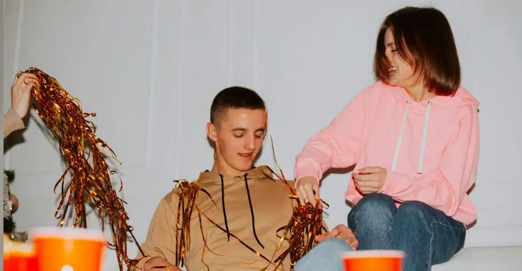 Two friends sitting on a couch, laughing and playing with gold streamers, surrounded by party decorations.