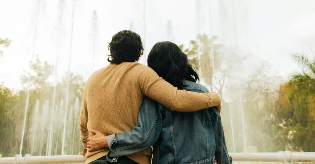 A couple with their arms around each other, standing in front of a tall fountain in an outdoor setting.