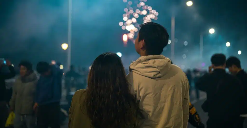 A couple standing among a crowd, looking up at fireworks in the night sky.