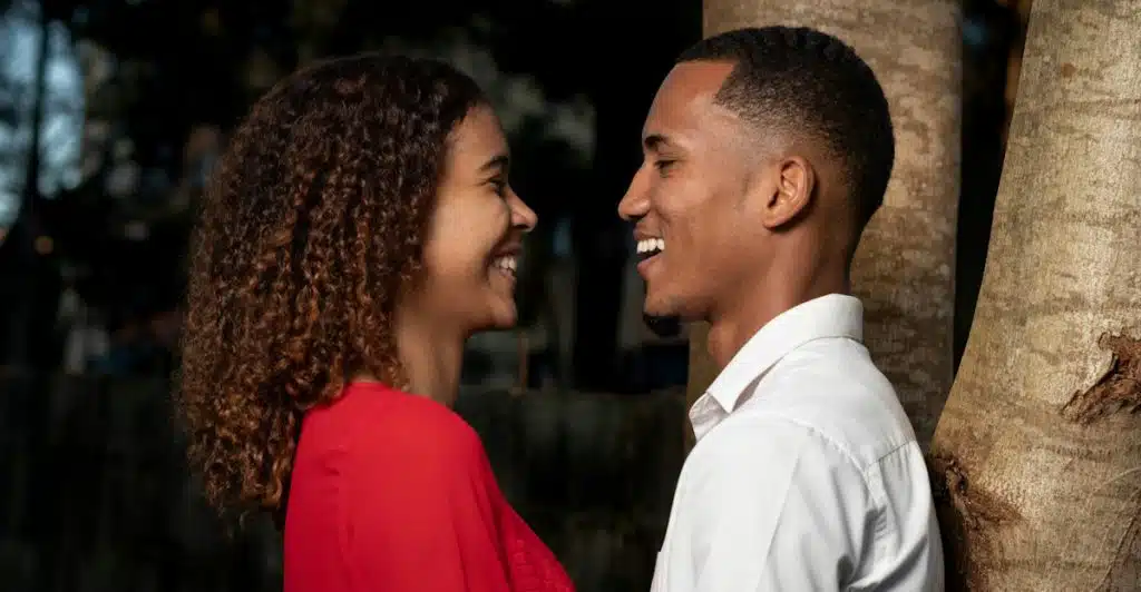 A couple standing close to each other, smiling and looking into each other's eyes, with trees in the background.
