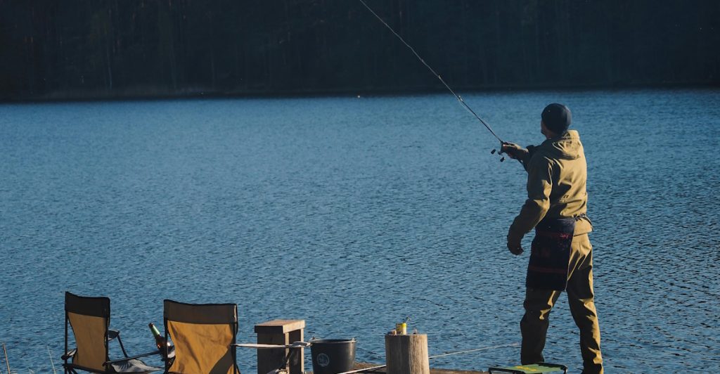 A man wearing a green jacket and a blue beanie fishing with a long rod on a wooden dock by a calm blue lake.