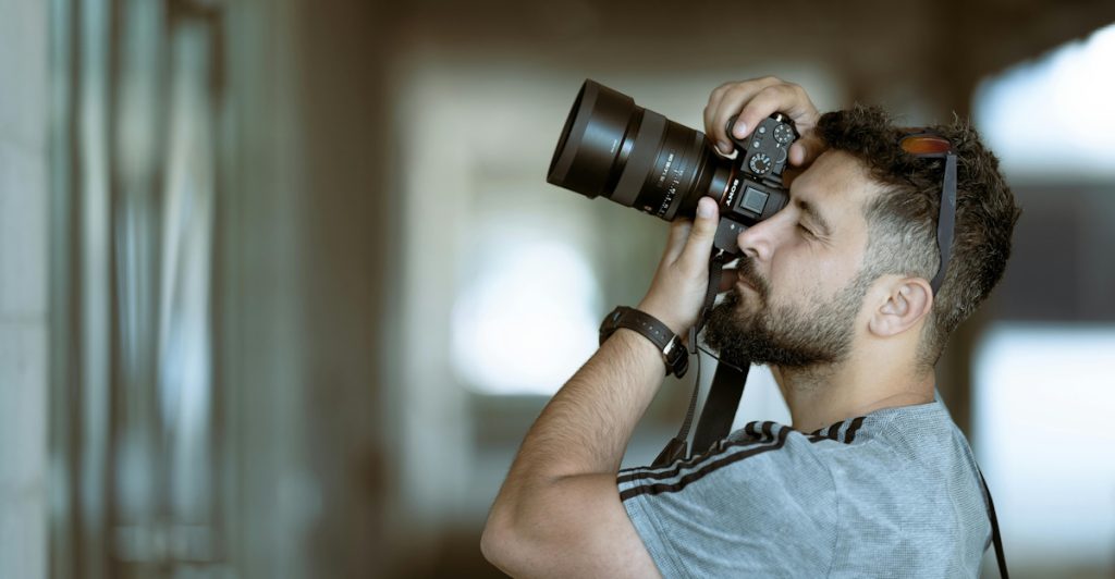 A bearded man wearing a gray t-shirt holding a camera with a large lens up to his eye, focusing on taking a photograph.