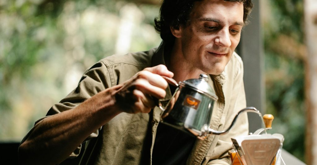A man wearing a light green shirt carefully pouring hot water from a small metal kettle into a coffee filter setup on an outdoor table.