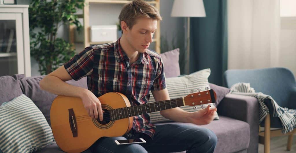 A man with blond hair wearing a checkered shirt sitting on a gray couch, tuning or playing an acoustic guitar.