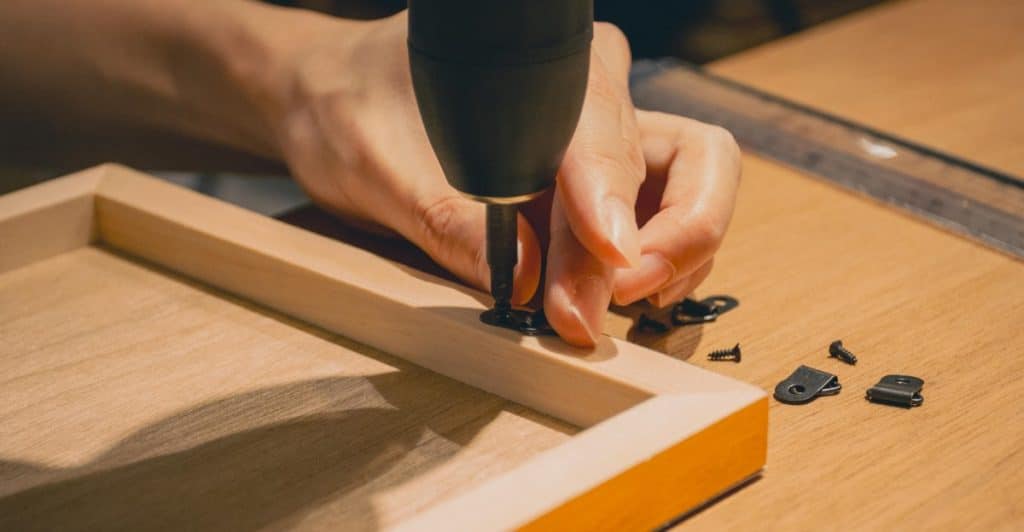 Close-up of hands using a drill to screw a small black bracket onto a wooden frame.