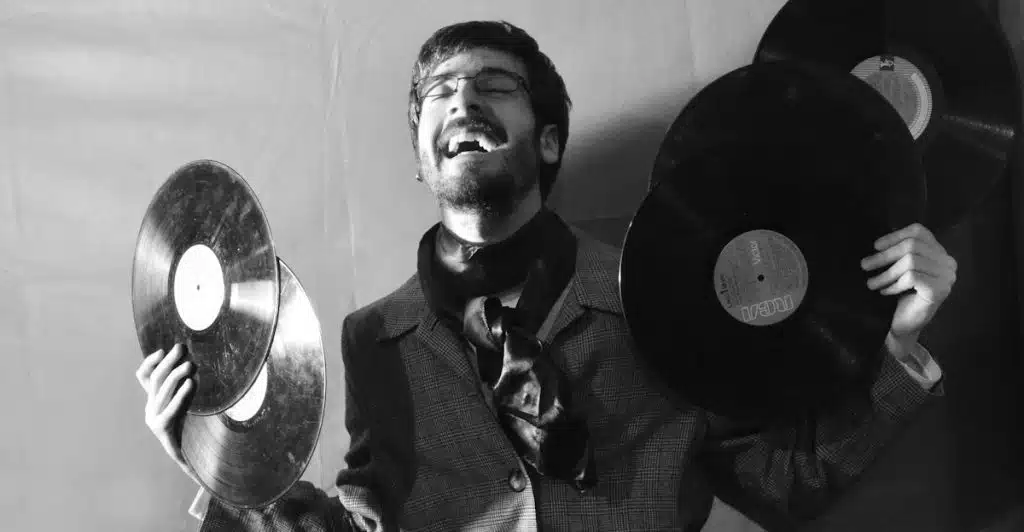 A man with glasses and a scarf laughing and holding up two vinyl records in a black and white photo.