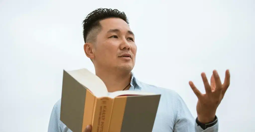 A man with short dark hair holding an open book and gesturing with his hand as if speaking or presenting.