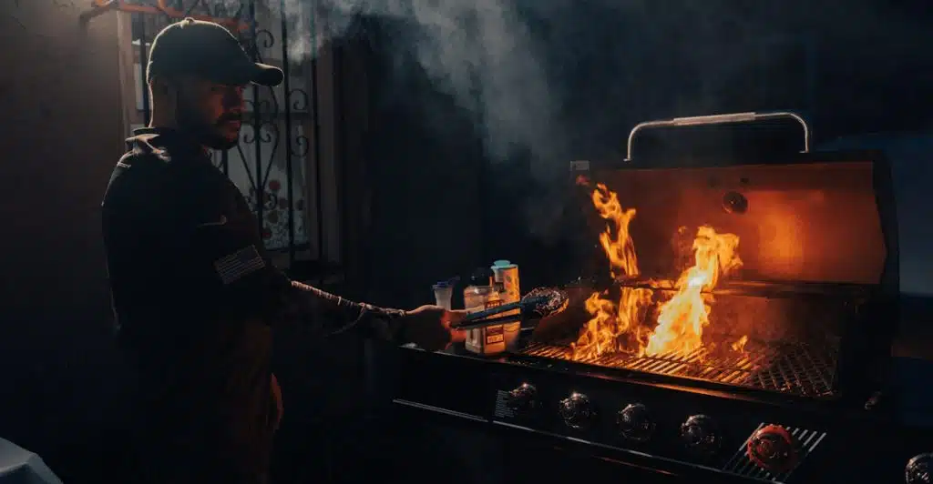 A man wearing a cap standing at a grill in the evening, tending to flames and smoke.
