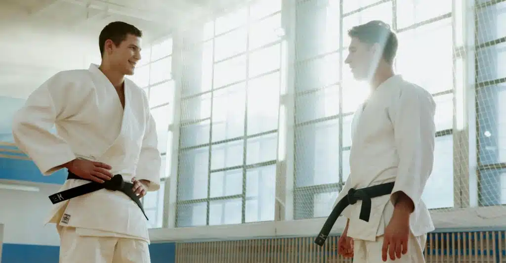 Two men wearing white martial arts uniforms with black belts facing each other in a training space with large windows.