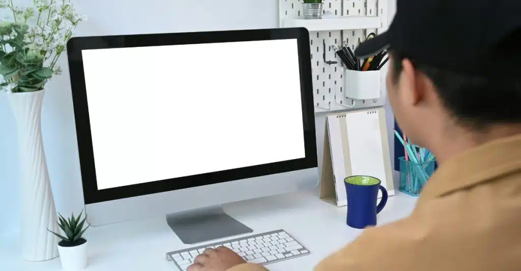 A person wearing a tan jacket and a black cap using a computer with a white screen, keyboard, and mouse on a white desk.