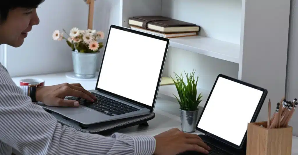 A person wearing a striped shirt using two laptops on a white desk with office supplies.