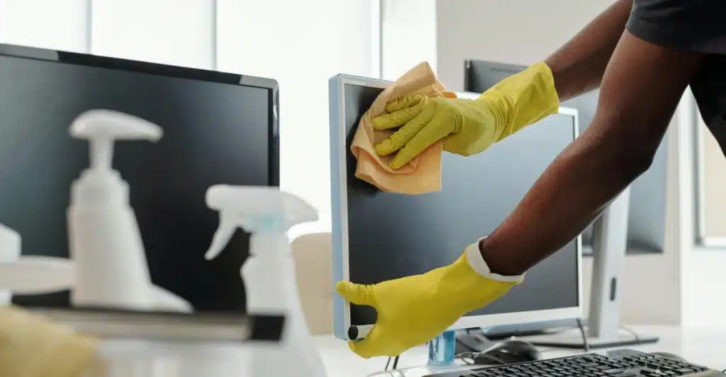 A person wearing yellow gloves cleaning computer screens with a cloth and cleaning spray bottles nearby.