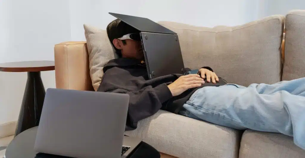 A person lying on a couch covering their eyes with a laptop, with another laptop on a nearby table.