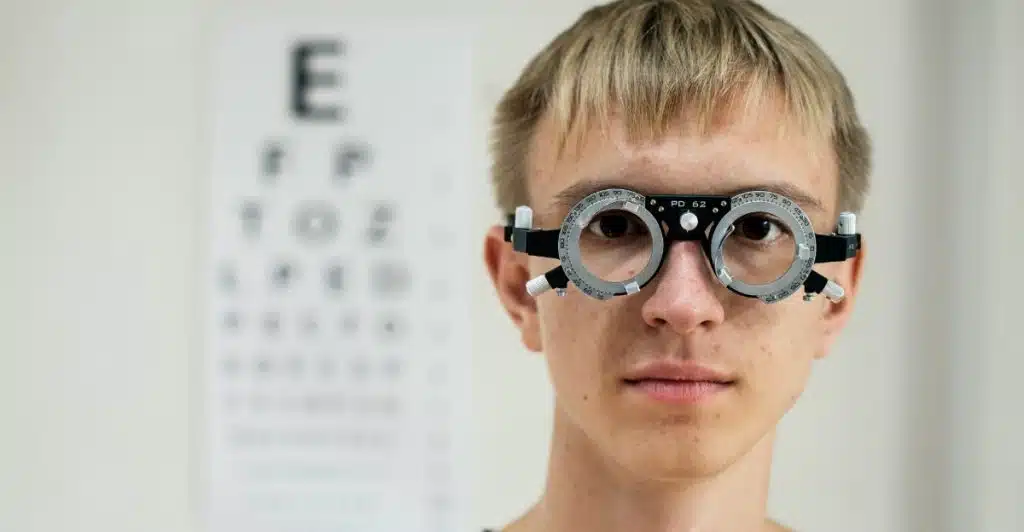 A person with light-colored hair wearing an eye testing apparatus with an eye chart in the background.