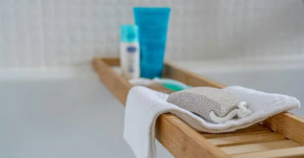 A wooden bath caddy rests on the edge of a white bathtub, holding a pumice stone, a white towel, and various blue and white toiletry tubes.