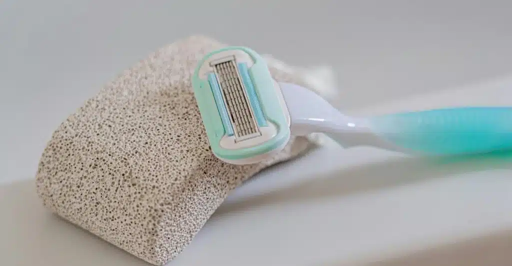A light blue and white razor leaning against a porous pumice stone on a white surface.