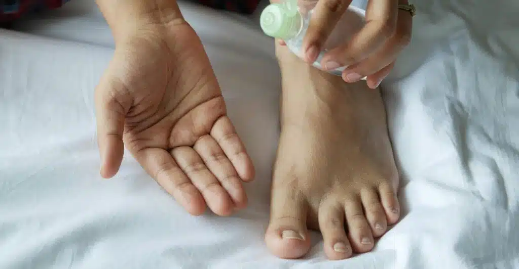 A person's hands applying lotion from a white bottle to their foot, resting on a white sheet.
