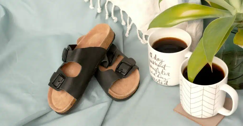 A pair of dark-colored sandals next to two mugs of coffee on a light blue surface, with a plant in the background.
