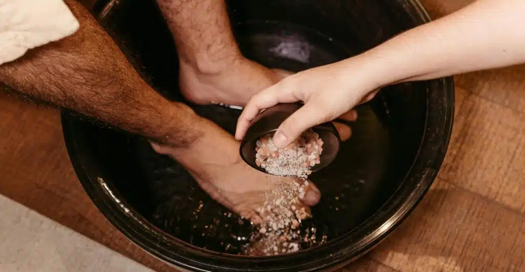People's feet soaking in a dark basin while another person pours a grainy substance from a small bowl.